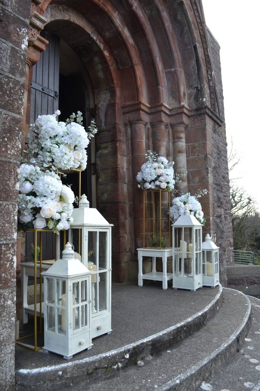 white Lanterns and floral display stands on steps and at either side of church entrance