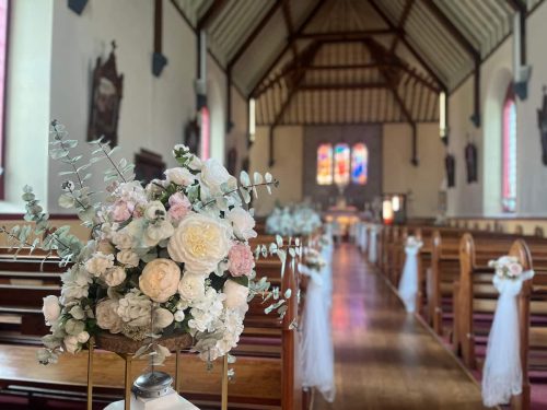 Close up of flower stand with church aisle in the background by Rosewater Decor