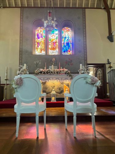 Wedding chairs with flowers in front of altar and stained glass window by Rosewater Decor